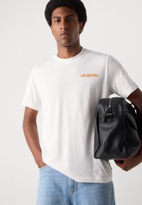 Weißes Baumwoll-T-Shirt mit dem bedruckten orangenen Text "DEIN ABENTEUER", kombiniert mit einem schwarzen Leder-Rucksack mit Reißverschluss.