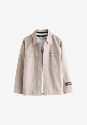 Teenagedreng iført en beige jakke med knapper og matchende bukser med "Baker"-branding på begge dele mod en ensfarvet baggrund.