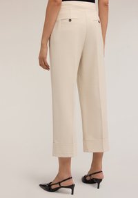 Pantaloni beige a gamba larga con una texture liscia, dotati di due tasche posteriori e orli risvoltati. Abbinati a scarpe con tacco nere.