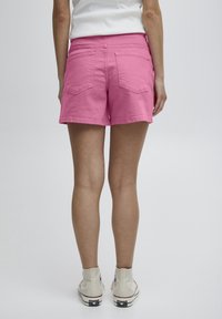 Pantalones cortos de algodón rosa con una cintura y dos bolsillos traseros, combinados con zapatillas blancas de caña alta, vistos desde atrás.