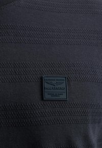 Camiseta a rayas negra hecha de tejido suave, con un patrón texturizado, escote redondeado y un parche de goma cuadrado en el pecho.