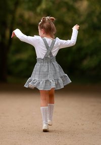 Młoda dziewczyna z kokardkami we włosach, ubrana w białą bluzkę, spódniczkę w kratkę z szelkami i skarpetki do kolan, spaceruje na świeżym powietrzu z uniesionymi rękami.