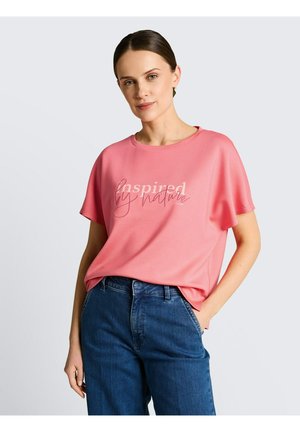 Kobieta w różowym t-shircie z napisem „inspired by nature” i niebieskich dżinsach, pozująca z jedną ręką w kieszeni na jednolitym tle.