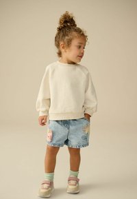 Felpa bianca in pile con polsini a costine abbinata a pantaloni shorts di jeans azzurri chiari con disegni ricamati di fiori e animali. Calzini verde chiaro e sneakers.