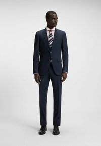 Granatowy garnitur z wyraźnymi ramionami, zapięciem na jeden guzik i klapkami. W połączeniu z jasnoróżową koszulą i wąskim krawatem w paski. Czarne buty.