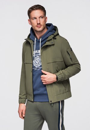 Hombre con chaqueta verde con capucha sobre sudadera azul con capucha y pantalones deportivos grises con rayas laterales, de pie sobre fondo liso.