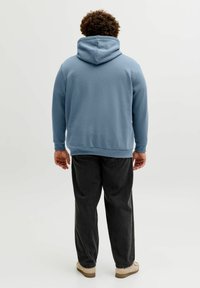 Modra jopa s kapuco s prednjim žepom in vrvico, združena s temno sivimi hlačami in bež čevlji, viden od zadaj.