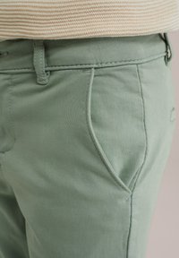 Svetlozeleni bombažni hlače imajo gladko teksturo, detajle sprednjih žepov in šivane šive na pasu ter straneh.