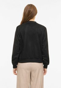 Juoda bombonešio striukė su lygiu zomšos paviršiumi, ribinėmis manžetėmis ir juosmens juostele. Matoma iš nugaros, be matomų aparatūros elementų ar raštų.