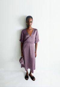 Lavender-colored dress with a V-neck, short rolled sleeves, and a flowing silhouette, paired with black slide sandals and a matching bag.