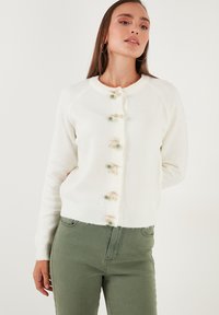 Cárdigan blanco de punto con cuello redondo, que presenta botones decorativos con acentos florales en beige y verde. Mangas largas, cintura ajustada.