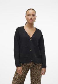 Schwarzer Cardigan mit V-Ausschnitt, vorderen Knöpfen, gerippten Kanten und zwei Fronttaschen. Weiche Textur, lässige Passform, kombiniert mit Hosen im Leopardenmuster.
