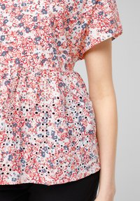 Blouse fleurie en tissu léger avec un imprimé multicolore de fleurs rouges et bleues, taille cintrée et détails œillets le long de l'ourlet.