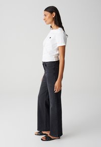 Witte katoenen T-shirt met een kleine zwarte hartafbeelding, gecombineerd met donkere denim wijde broek en zwarte platte sandalen met banden.