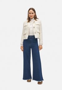 Chaqueta corta blanca texturizada con grandes bolsillos, combinada con unos vaqueros de denim azul de pierna ancha de cintura alta y zapatos de plataforma con estampado animal.