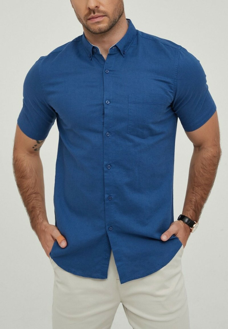 Chemise à manches courtes avec boutonnage en bleu profond, dotée d'une poche poitrine, d'une texture lisse, d'un col structuré et de détails subtils sur les boutons.