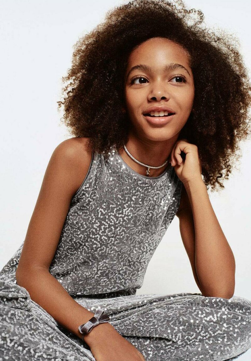 Srebrna bleščeča obleka brez rokavov z mehko žametno teksturo in usklajeno zapestnico. Model ima kodraste lase in naraven izraz.