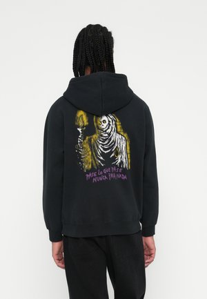 Crna hoodie sa grafikom skeletne figure koja drži svijeću, s žutim i ljubičastim tekstom na leđima. Materijal izgleda mekan s opuštenim krojem.