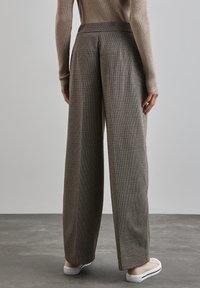 Korkeavyötäröiset, leveälahkeiset housut ruskeassa houndstooth-kuosissa, joissa on litteä etuosa ja taitteet lahkeissa, yhdistettynä kermaisiin lenkkitossuihin.