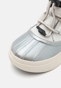 Zapatilla plateada con un acabado texturizado, paneles lisos y suela de goma blanca. Presenta cordones elásticos y una forma de punta redondeada.