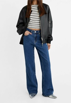 Kvinde iført sort bomberjakke, sort-hvidstribet crop top, viddeblå jeans og sølvspidse sko.