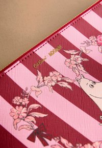Sarkans un rozā svītrots tekstils maks ar ziedu rakstiem, kurā ir Moomin ilustrācija un zelta zīmogs. Redzama teksturēta virsma un šuves.