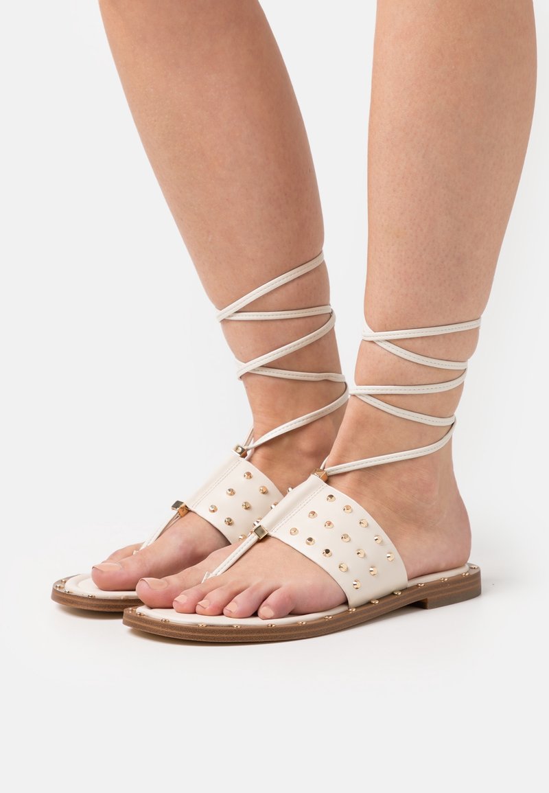 Sandali di pelle bianca con borchie dorate, design open toe, suola piatta e cinturini alla caviglia avvolti attorno alle gambe per un supporto aggiuntivo.