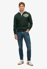Donkergroene pullover met contrasterende witte kraag, met het "SUPER DRY" logo in beige; gecombineerd met blauwe jeans en witte sneakers.