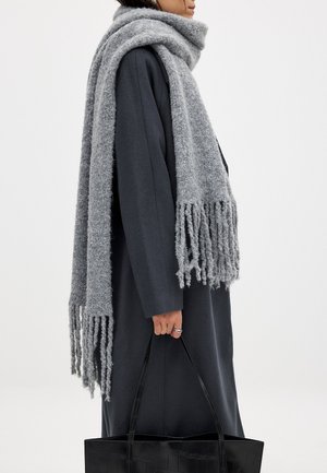 Écharpe en laine grise avec franges, drapée sur un manteau sombre. La personne tient un sac fourre-tout noir et brillant dans une main. La texture de l'écharpe semble douce et duveteuse.
