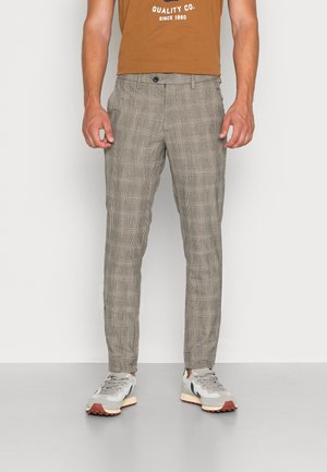 Mann trägt beige karierte, schmal geschnittene Hosen, weiße Sneaker und ein braunes T-Shirt mit weißem Text, steht vor neutralem Hintergrund.