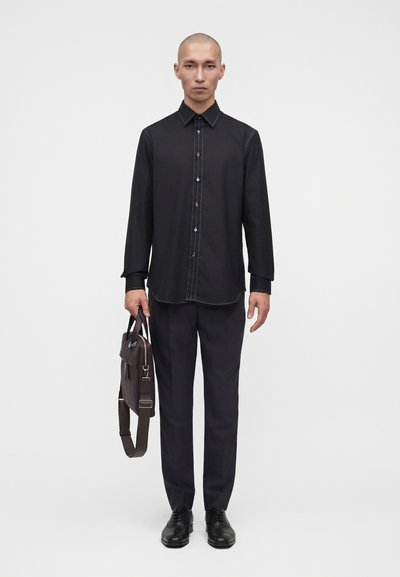 Homme debout portant une chemise noire boutonnée, un pantalon noir, des chaussures élégantes noires, tenant une mallette marron foncé sur un fond blanc uni.