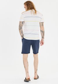 Miestn1 a kr1kym rukavom, pruhovan3 tri1ko v bielej farbe s viacermi farebnmi horizontlnymi pruhmi, skombinovan1 s navy kraťasmi. Na nahj nohe s nosn1 sand0ly.