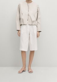 Person iført en beige lynlåstrøje, hvide knælange stribede shorts og hvide sandalholder mod en ensfarvet baggrund.