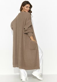 Długi, gruby, dziany kardigan w jasnobrązowym kolorze, z oversize'owymi rękawami, luźnym krojem i kieszeniami z przodu. Teksturowana tkanina dodaje głębi.