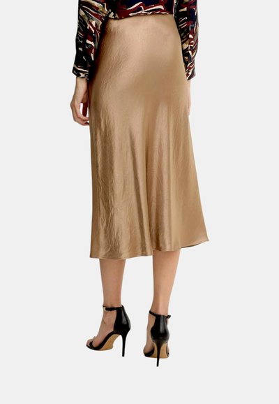 Donna che indossa una gonna midi beige lucida, una maglia a maniche lunghe con motivo e sandali neri con tacco alto e cinturino alla caviglia, in piedi con le mani rilassate.