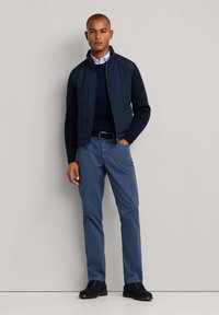 Granatowa pikowana kamizelka noszona na granatowym swetrze, zestawiona z jasnoniebieskimi spodniami. Czarny pasek i ciemne buty dopełniają całości. Gładkie tekstury materiałów.