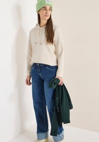 Femme portant un bonnet vert, un sweat à capuche beige, un jean bleu retroussé, tenant une veste boutonnée vert foncé, debout contre un mur blanc.
