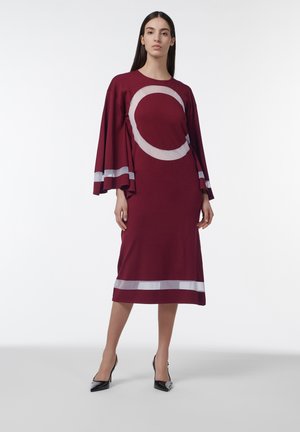 Femme debout portant une robe midi bordeaux à larges manches et motifs grands cercles pâles et rayures, associée à des escarpins noirs pointus.