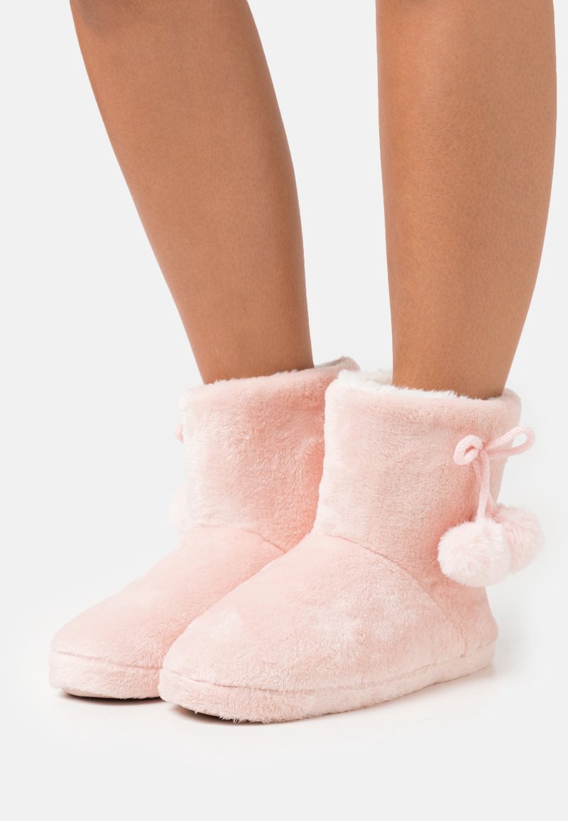 Bottes basses roses en peluche avec une texture douce, dotées d'un design replié et d'accent pompons sur le côté, adaptées à une utilisation en intérieur.