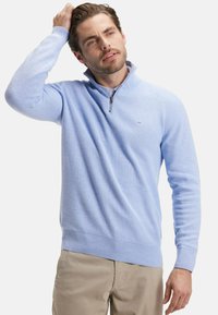 Pull zippé bleu clair en tissu tricoté texturé avec des poignets et un ourlet côtelés. Détail logo discret sur la poitrine. Pantalon de couleur neutre.