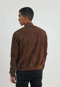 Veste bomber en cuir suedé marron avec des poignets et un ourlet côtelés, présentant une étiquette de marque discrète à l'arrière et une texture lisse et douce.
