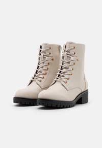 Botines de piel beige con ojales dorados, cordones y cremallera lateral; puntera redondeada y suela negra gruesa con relieves para mayor tracción.