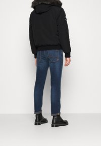 Man wearing dark blue rolled-up jeans, black leather boots, and a black jacket with a fur-trimmed hood, standing against a white background.