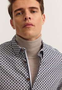 Koszulka button-up w wzory w odcieniach bieli, niebieskiego i żółtego, noszona na jasnym beżowym golfie. Posiada kołnierz rozłożony i brązowe guziki.