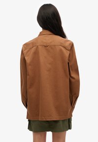 Brązowa, oversize'owa kurtka bawełniana z kołnierzem, długimi rękawami i bocznymi rozcięciami. Materiał ma gładką teksturę i jednolity wzór.