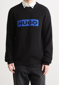Schwarzer Strickpullover mit gerippter Textur, versehen mit einem blauen rechteckigen Patch, auf dem das Wort "HUGO" in fetten schwarzen Buchstaben über der Brust steht.