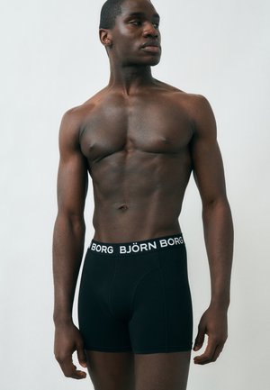 Homme en forme torse nu portant un boxer Björn Borg noir, debout devant un fond clair uni, regardant sur le côté.