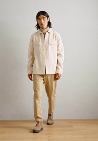 Light beige button-up shirt with two chest pockets, paired with tan trousers and beige boots. Smooth fabric and relaxed fit.