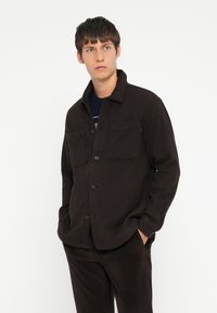 Black button-up jacket with a collar, two chest pockets, and a relaxed fit. Material appears textured and fabric looks durable.