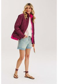 Prešívaná burgundská bunda s jasne ružovým podšívkou, nosená cez svetlé tričko s grafickým motívom a svetlomodré denimové šortky, doplnená hnedými sandálmi.